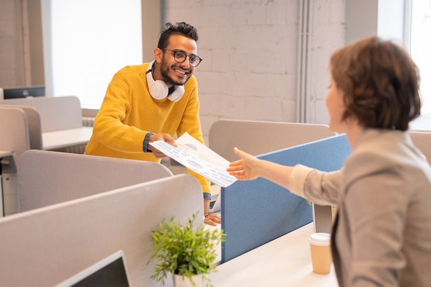 Positieve Arabische man met koptelefoon op nek staan aan bureau in open ruimte kantoor en financiële papieren geven aan collega