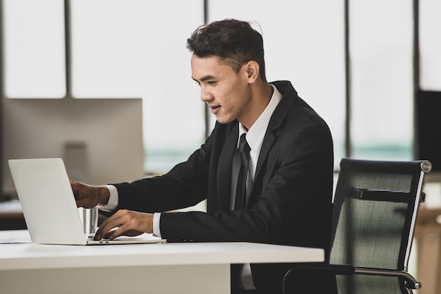 Positieve Aziatische mannelijke ondernemer die aan tafel zit en netbook bladert terwijl hij aan een project op de werkplek werkt en op zoek is naar een laptopcomputerscherm.