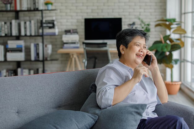 Positieve Aziatische vrouw van middelbare leeftijd die lacht en wegkijkt terwijl ze op de bank zit en een smartphonegesprek voert in de moderne woonkamer thuis