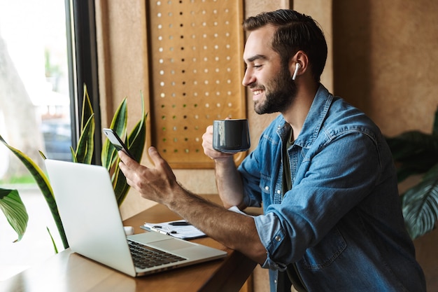 positieve bebaarde man met een denim shirt met een kopje thee en een smartphone terwijl hij binnenshuis in een café werkt