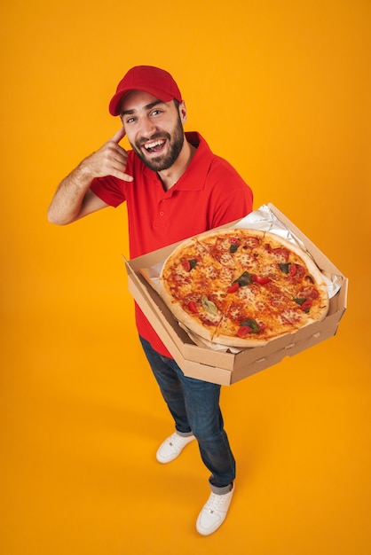 Foto positieve bezorger van volledige lengte in rood uniform glimlachend en met pizzadoos geïsoleerd over geel