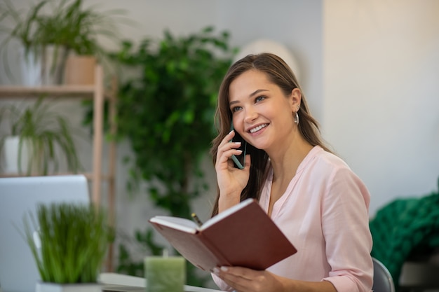 Positieve blije onderneemster die haar notitieboekje houdt tijdens het voeren van een telefoongesprek