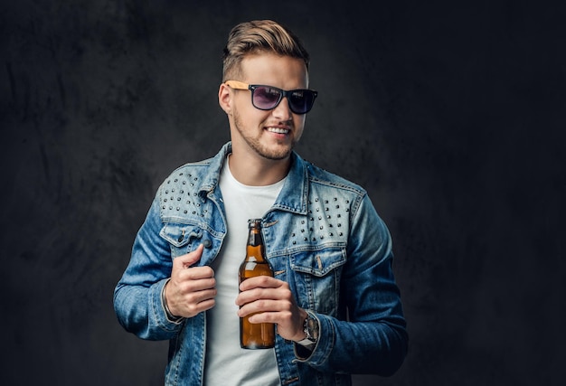 Positieve blonde man gekleed in een spijkerjasje en zonnebril houdt een fles met ambachtelijk bier vast.