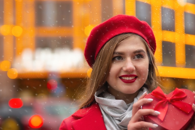 Positieve blonde vrouw draagt een rode baret en jas met een hartvormige geschenkdoos op de achtergrond van bokehlichten. Ruimte voor tekst