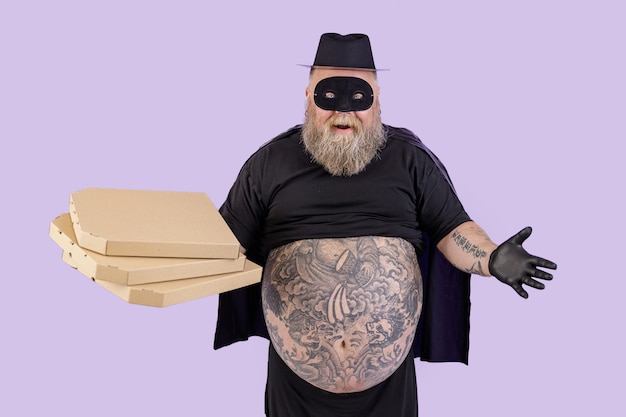 Foto positieve dikke man in zorrokostuum houdt kartonnen dozen pizza vast op paarse achtergrond