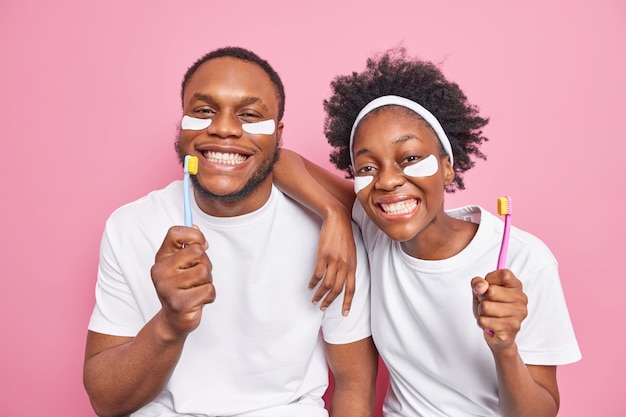Positieve donkere huid Afro-Amerikaanse vrouw en man glimlach hebben in grote lijnen perfect wit, zelfs tanden zorgen voor mondhygiëne, houden tandenborstels, brengen schoonheidspleisters aan geïsoleerd over roze muur