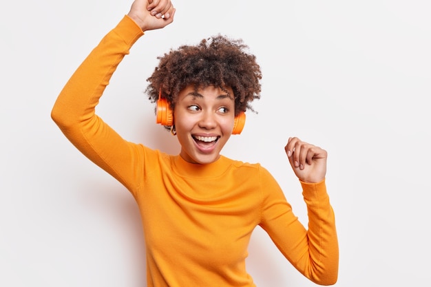 Positieve donkere jonge vrouw met krullend haar heft armen op en danst zorgeloos draagt casual jumper glimlacht breed vangt elk stukje muziek beweegt met ritme geïsoleerd over witte muur