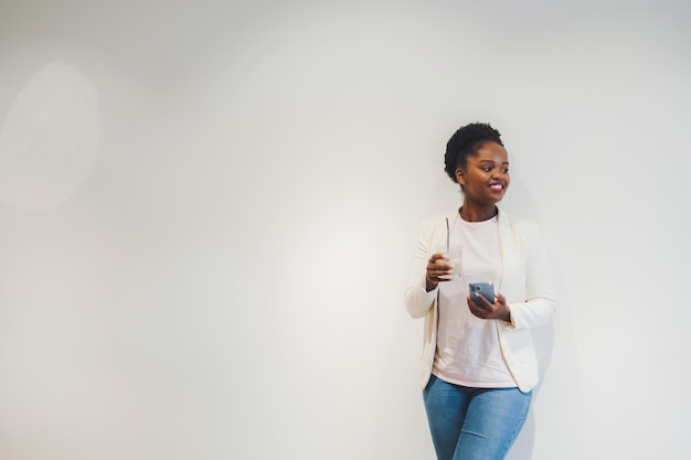 Foto positieve donkere vrouw geniet van werkpauze in coffeeshop, drinkt koude dranken en sms't berichten op mobiele telefoon leunend tegen de witte muur met vrije ruimte voor tekst kopieer ruimte