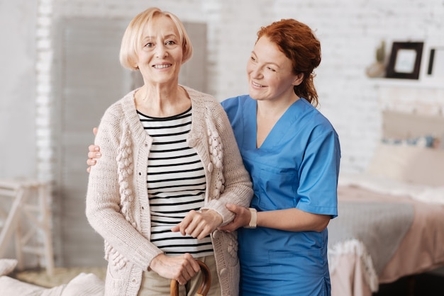 Positieve emoties. Bekwame, ondersteunende, zorgvuldige dame die de oudere vrouw bij haar handen vasthoudt en haar helpt een wandeling door haar appartement te maken