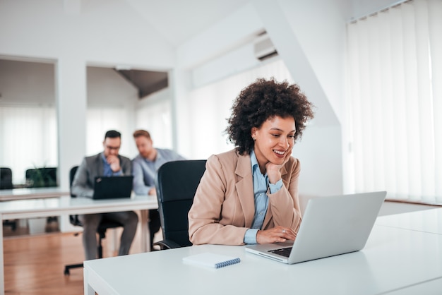 Positieve exotische onderneemster die aan laptop in gedeeld bureau werkt.