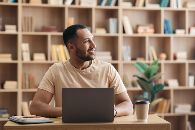 Positieve freelance man aan het werk op laptop thuis zittend op de werkplek en opzij kijkend naar kopie