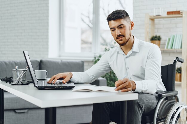 Positieve gehandicapte jonge man in rolstoel werken in kantoor met laptop