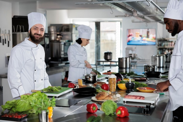 Positieve glimlachende chef-kok in de professionele keukenruimte van het restaurant die gastronomische maaltijd bereidt met behulp van heerlijke ingrediënten. Culinaire servicemedewerker die salade snijdt terwijl hij naar de camera kijkt.