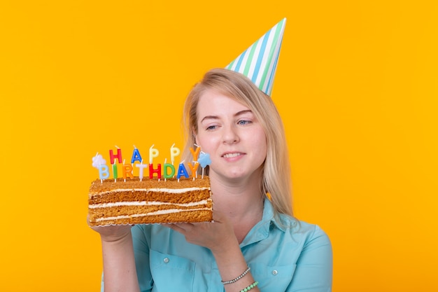 Positieve grappige jonge vrouw met een pet en een zelfgemaakte cake in haar handen die zich voordeed op een gele ondergrond. Verjaardag en verjaardag concept.