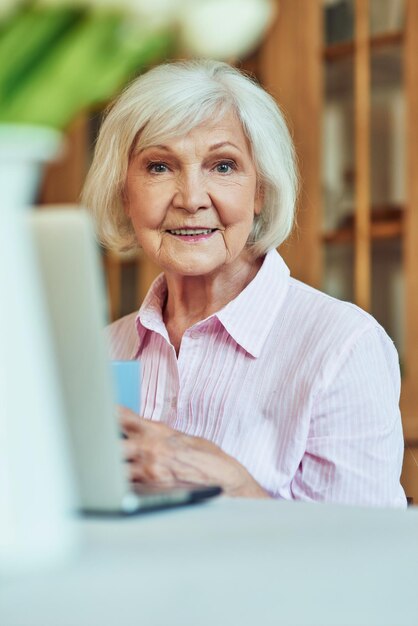 Positieve grijsharige dame die met de computer in de kamer werkt