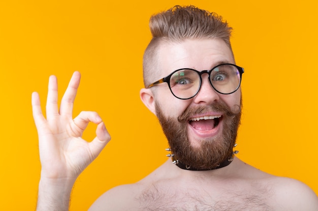 Positieve hipster shirtless man met een rocker-kraag toont ok op een gele muur. Concept ongebruikelijke groeten.