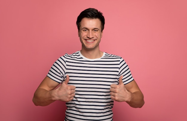 Positieve instelling. jonge man in een gestreept t-shirt, die in de camera kijkt, glimlachend en duimen opdagen.