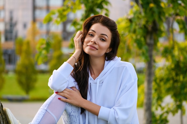 Positieve jonge brunette vrouw op de achtergrond van het groene stadspark na uitrekken, fitness in witte hoodie en leggings