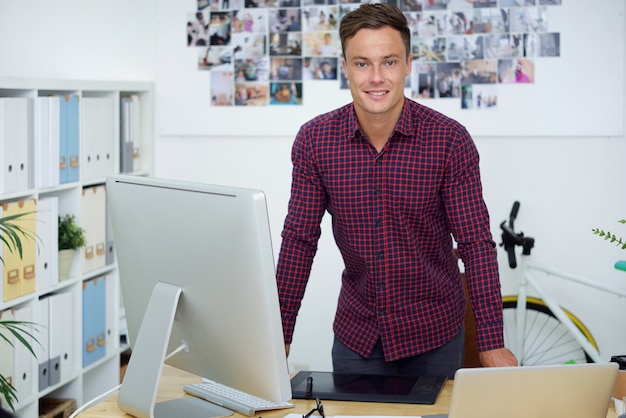 Positieve jonge grafisch ontwerper of fotograaf die aan zijn bureau staat met computer en grafisch tablet