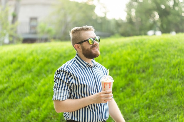 Positieve jonge hipster-man met een bril en een stijlvolle baard drinkt een milkshake met een rietje terwijl hij buiten tegen een bakstenen muur staat. Het concept van liefhebbers van lichtzoete dranken