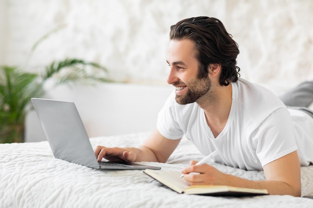 Positieve jonge man die op bed ligt met behulp van laptop die aantekeningen maakt