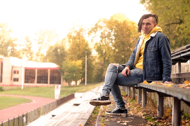 Positieve jonge man met dreadlocks kijkt naar de camera op de oude, armoedige bank van de straatbleker in de gouden herfst