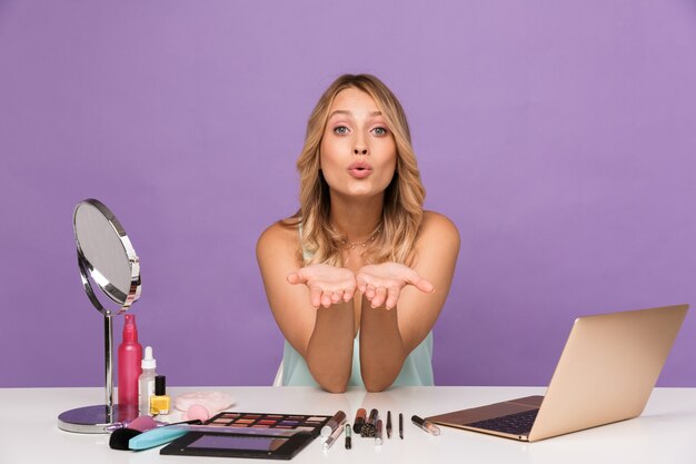 positieve jonge mooie vrouw met laptopcomputer en cosmetica met spiegel blazende kusjes.