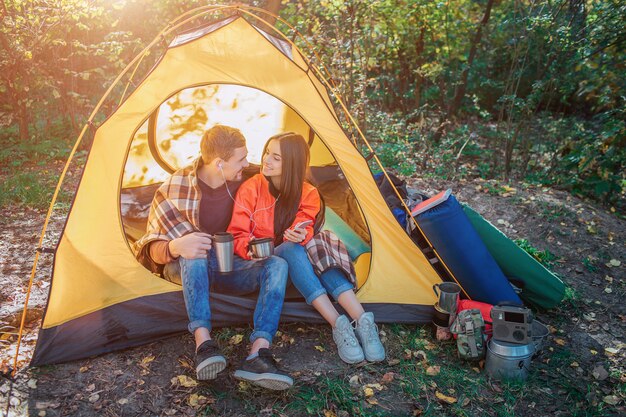 Positieve jonge paarzitting in tent en luister naar muziek door één hoofdtelefoon. ze kijken elkaar aan en glimlachen. ze houdt de telefoon vast. hij heeft thermosfles.