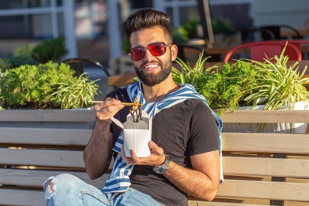 Positieve jonge stijlvolle Arabische man die Chinese noedels eet in een park tijdens een pauze op het werk. Het concept van rust en gezonde snack.