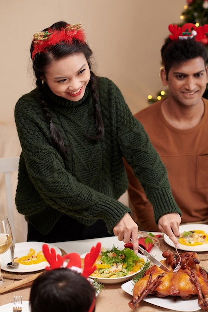 Positieve jonge vrouw die gebakken kalkoen snijdt voor gasten tijdens het kerstdiner