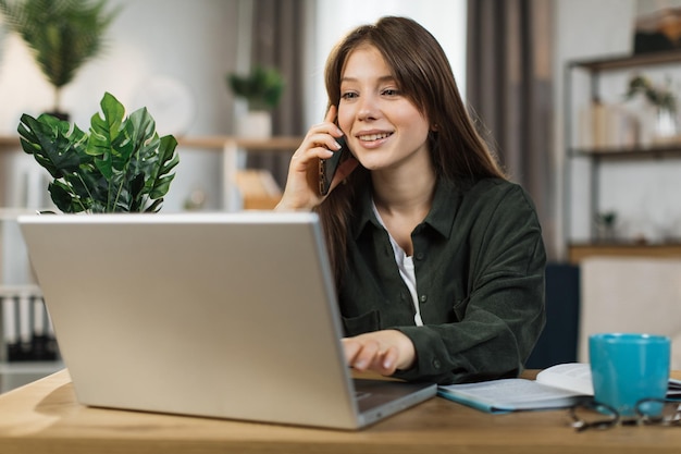 Positieve jonge vrouw die op mobiel praat en op een laptop werkt op een helder kantoor