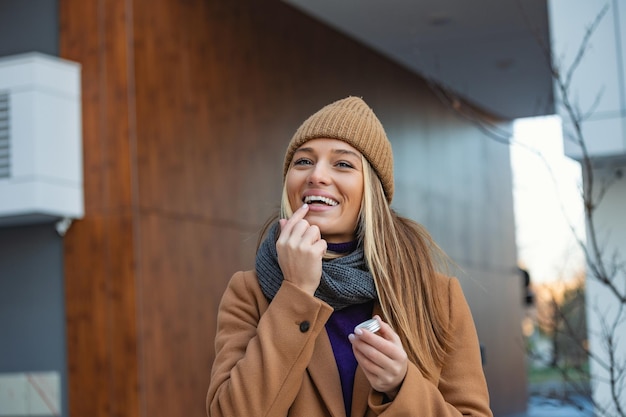 Positieve jonge vrouw in warme jas glimlachend en wegkijkend terwijl ze lippenbalsem op de lippen smeert tijdens een wandeling door de stad