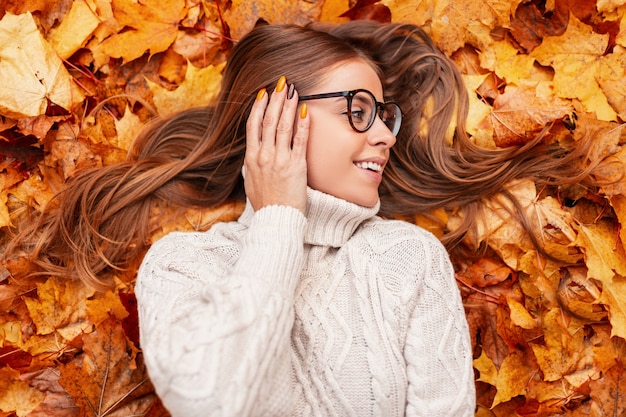 Positieve jonge vrouw met een mooie glimlach in een stijlvolle bril in een trendy witte gebreide trui rust in de herfst oranje gebladerte in het bos. Vrolijk meisje ontspant buiten in een park.