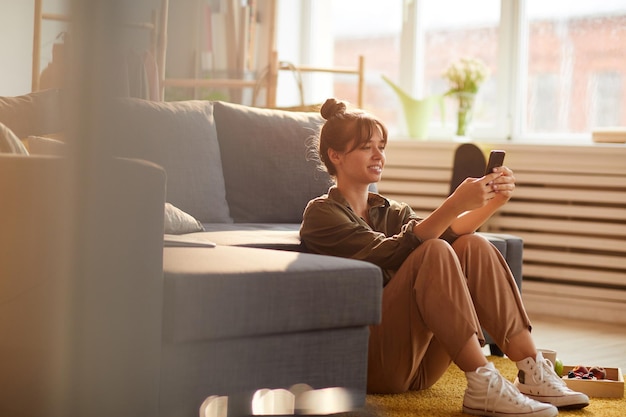 Positieve jonge vrouw met haarknot die op de vloer zit en op de bank leunt terwijl ze thuis een smartphone gebruikt