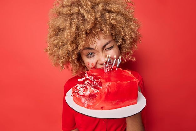 Positieve jonge vrouw met krullend haar knipoogt oog eet smakelijke slagroomtaart heeft gelekt make-up geniet van het eten van zoete dessert poses binnen geïsoleerd over levendige rode achtergrond geeft niet om calorieën