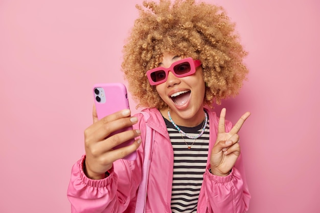 Positieve jonge vrouw met krullend haar vrouw maakt vredesgebaar lacht neemt graag selfie via smartphone draagt trendy zonnebril en jas geïsoleerd over roze achtergrond heeft plezier maakt foto