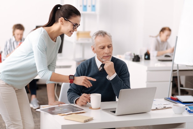 Positieve jonge vrouwelijke werknemer die slimme horloges draagt, leunt met haar linkerhand op de tafel wijzend op de computer