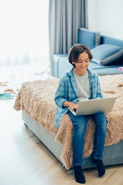 Positieve jongen in vrijetijdskleding die thuis ontspant en glimlacht terwijl hij naar het scherm van een laptop op zijn schoot kijkt