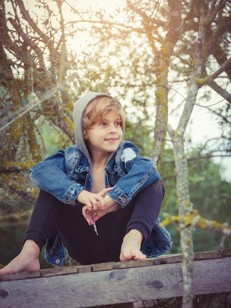 Positieve jongen op blote voeten in spijkerjasje met capuchon glimlachend en wegkijkend terwijl hij op een houtpad zit te midden van bladerloze boomtakken in het bos