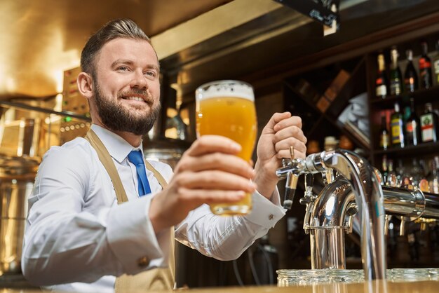 Positieve, knappe barman die een koud lagerbierglas houdt. Vrolijke brouwerij werknemer permanent aan toog, glimlachend. Volwassen man in wit overhemd en bruine schort camera kijken.