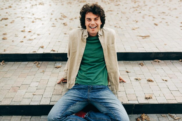 Foto positieve knappe jonge man zittend op de trap glimlachend en kijkend naar de camera outdoor portret van gelukkig slimme student man rust buiten mensen en emotie concept