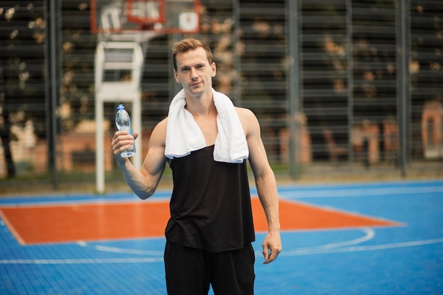 Positieve knappe man in sportkleding die zich voordeed op een basketbalveld buiten met een fles water