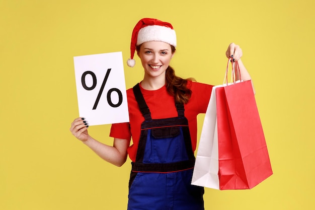 Positieve koeriersvrouw toont papier met procentteken, toont boodschappentassen, kortingen voor bezorgen, blauwe overalls en kerstmanhoed dragen. Indoor studio opname geïsoleerd op gele achtergrond.