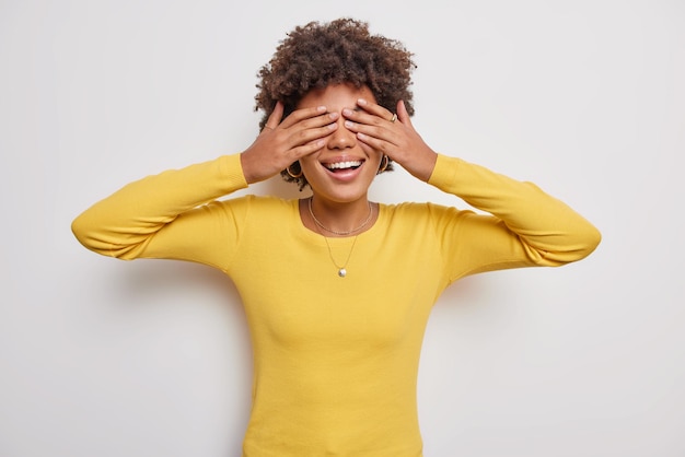 Positieve krullend haired jonge vrouw speelt verstoppertje dekt ogen met palmen glimlacht wacht op verrassing draagt gele trui geïsoleerd op witte achtergrond Blij vrouwelijk model maakt blind gebaar