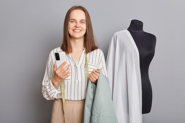 Positieve lachende optimistische vrouw kleermaker staande in de buurt van nieuwe jurk op etalagepop geïsoleerd over grijze achtergrond met slimme telefoon kijkend naar de camera met een blij gezicht