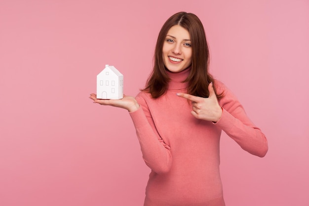Positieve lachende vrouw met bruin haar in roze trui wijzende vinger naar papier speelgoed huis in haar hand appartementen huur en verkoop makelaar binnen studio opname geïsoleerd op roze achtergrond