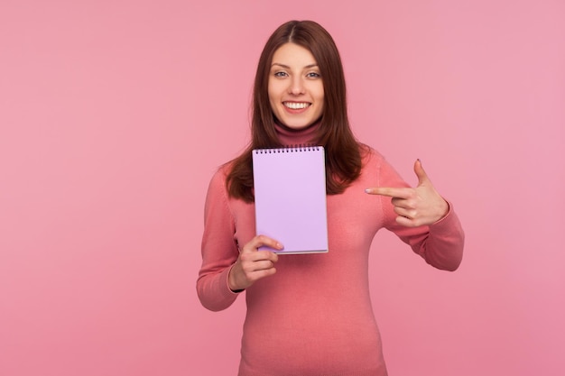 Positieve lachende vrouw wijzende vinger met notitieboekje met lege ruimte voor commerciële tekst, reclamegebied op papier voor plannen. Indoor studio-opname geïsoleerd op roze background