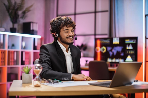Positieve man in headset met videogesprek met partners tijdens het werkproces 's avonds op kantoor