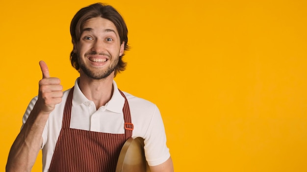 Positieve mannelijke ober met schort die duim omhoog houdt, keurde iets goed bij camera over oranje achtergrond Lichaamstaal