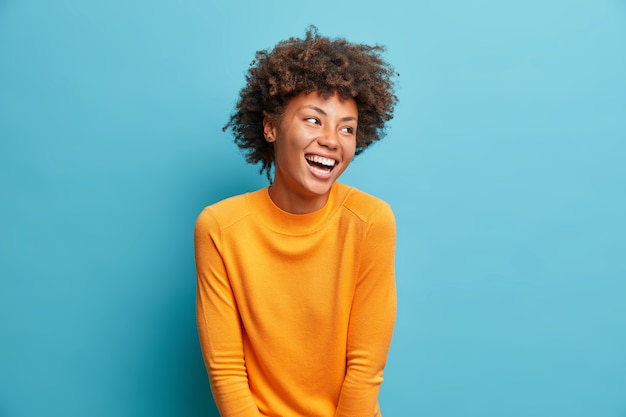 Foto positieve mooie jonge vrouw lacht giftig kijkt opzij met zorgeloze gezichtsuitdrukking draagt casual oranje trui geïsoleerd over blauwe studiomuur